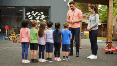 Two adults stand facing a group of young children lined up outside near playground equipment at a daycare or school.